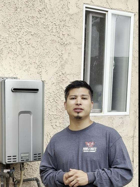 A Power Route technician in a branded shirt beside a tankless water heater installation.